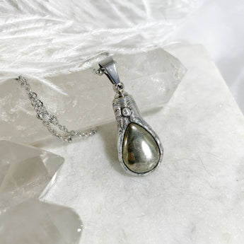 A silver necklace with a teardrop-shaped urn pendant. The pendant is filled with a gold-colored stone and has a detailed design.