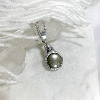 A silver urn necklace with a pendant shaped like a hand holding a round, pyrite stone. The stone is set in a silver frame and has a textured surface.